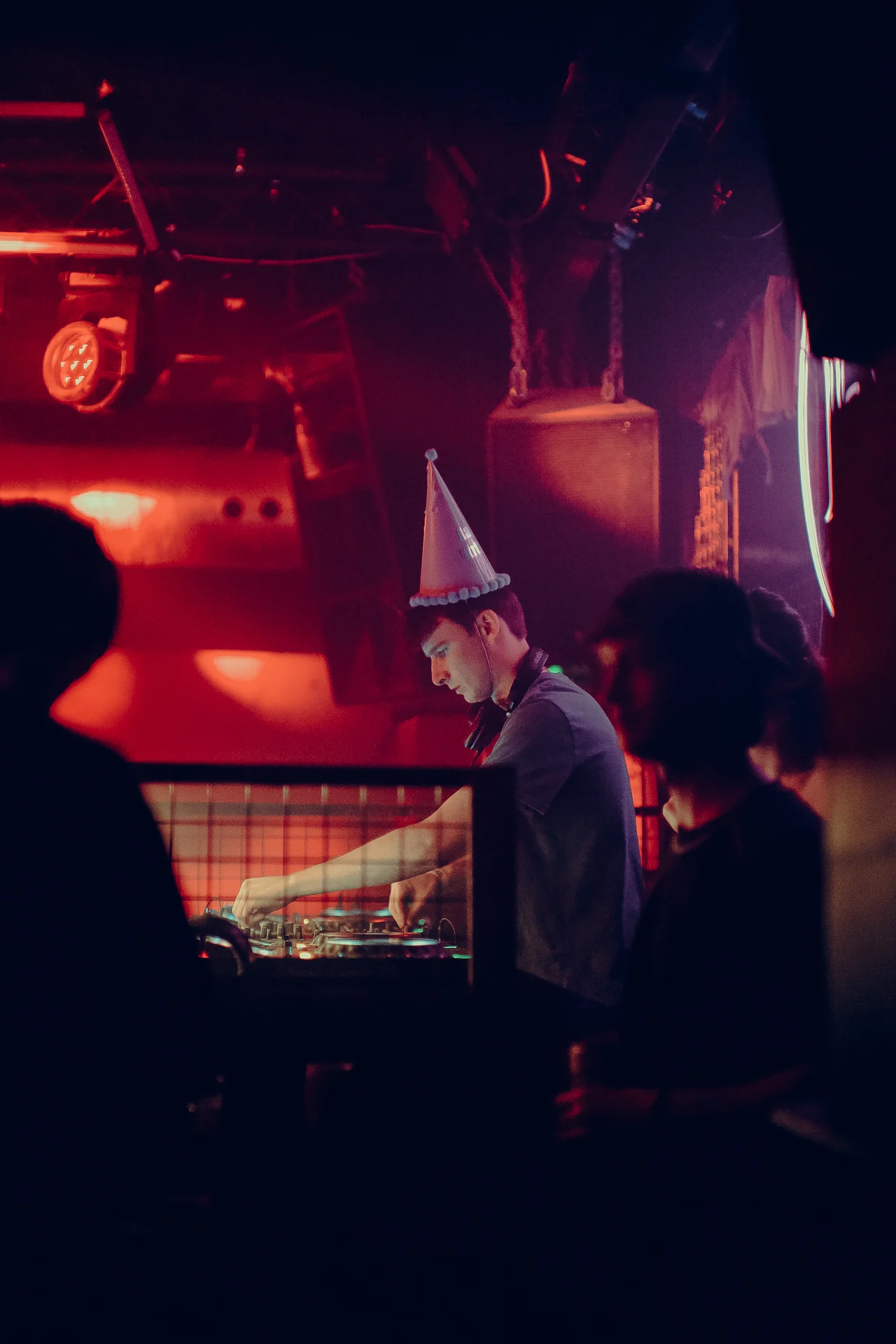 A DJ in a pointed party hat at the decks, framed by red light and silhouettes.