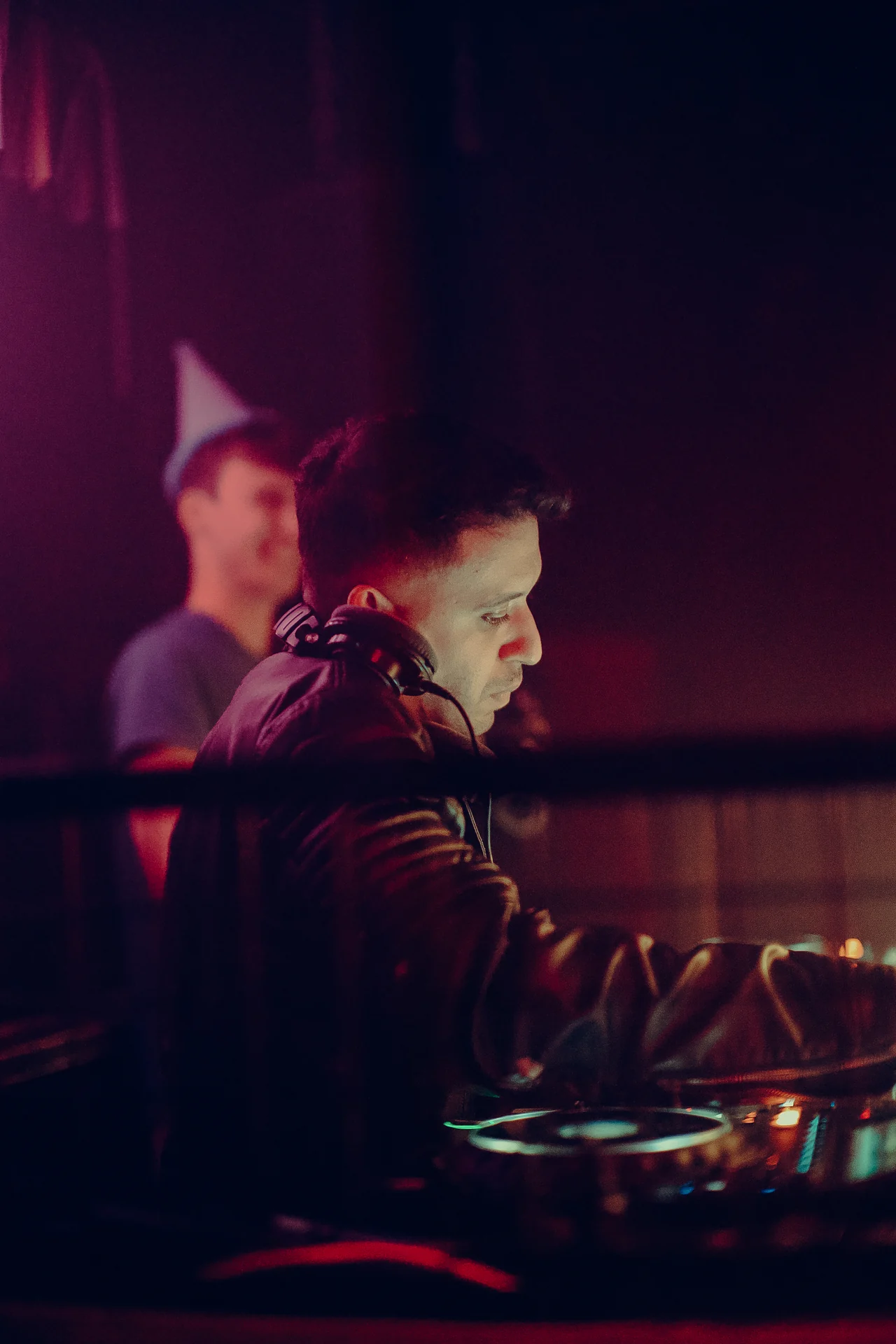 A DJ in a leather jacket leans into the mixer under red and violet light.
