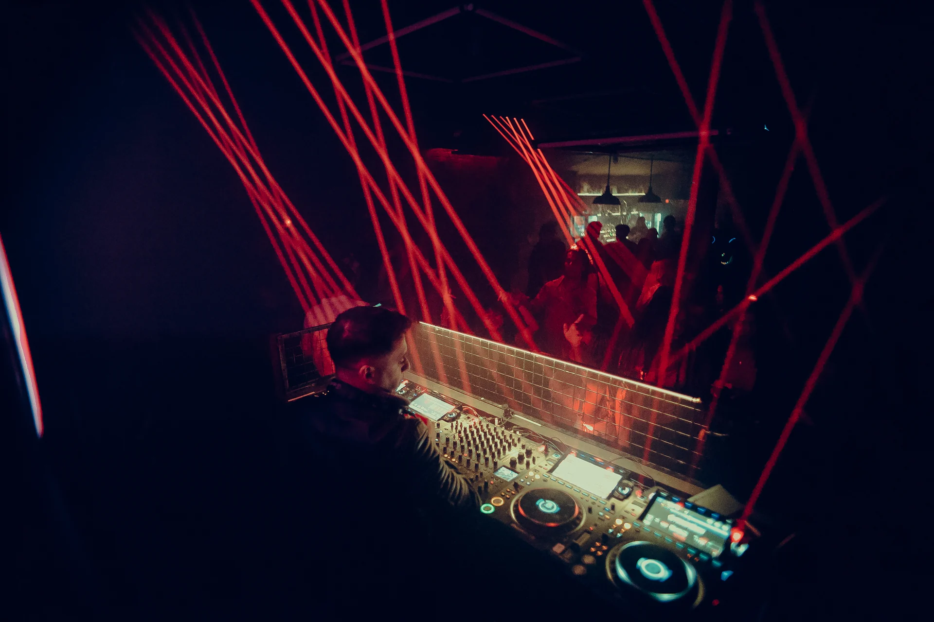 Red lasers fan over the decks and crowd in a wide view from behind the booth.