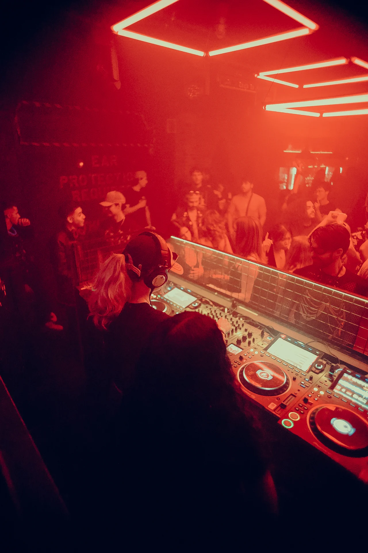 A DJ in headphones faces the red-lit dancefloor beneath geometric ceiling lights.