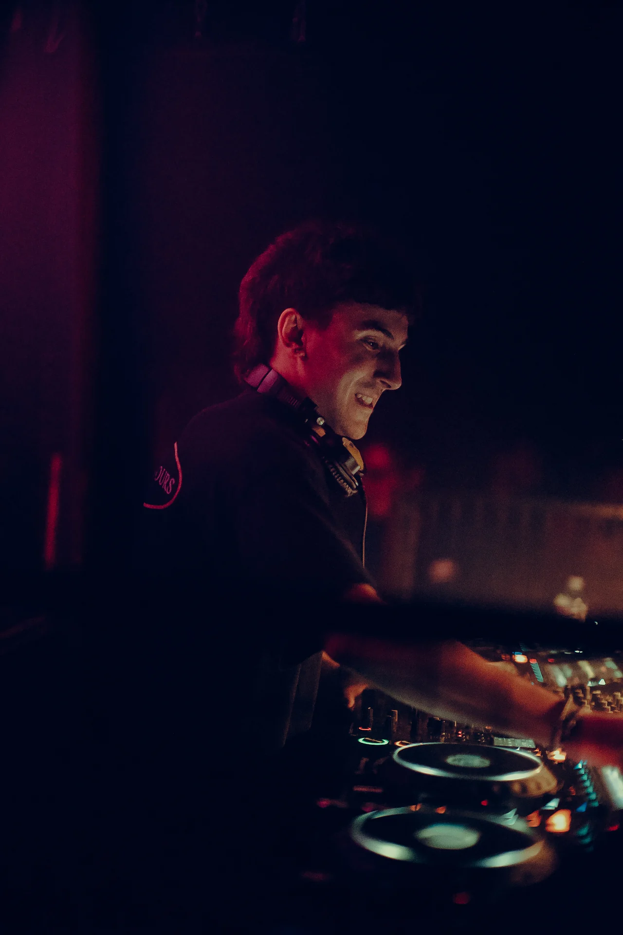 A smiling DJ reaches across the decks against a deep red and blue background.