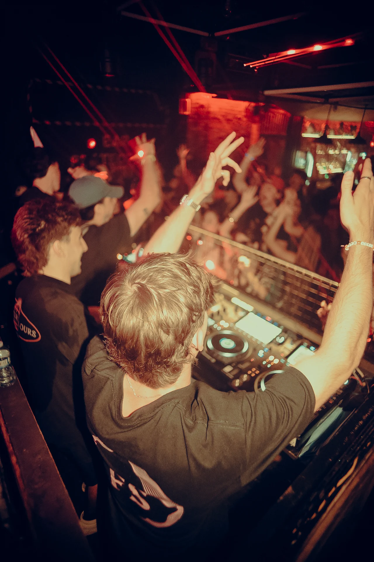 A DJ raises both arms over the decks as the front row responds under red lights.